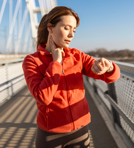a middle age woman woman wearing a red jacking standing on a bridge checking her pulse on her neck while she exercises